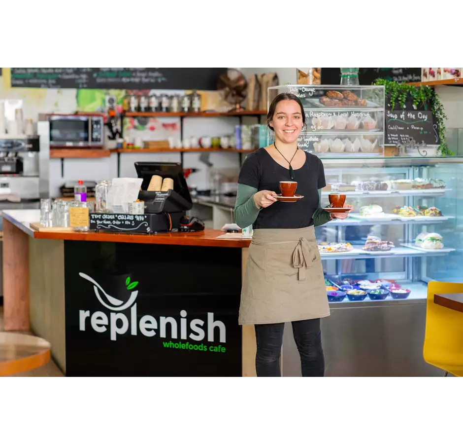 Replenish Cafe waitress holding two coffee cups in front of counter of food.